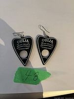 Photo of a pair of black Ouija board planchette-shaped earrings with white design and metal hooks on a white surface.