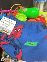 Blue Spiderman drawstring bag partially filled with assorted beach toys including a green water sprayer and orange handled items.