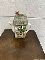 Front view of the ceramic teapot showing the cottage design with door, window, and foliage details.