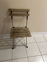 Front angled view of IKEA folding chair showing wooden slatted back and seat with black metal frame against tiled floor and beige wall.