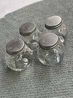 Four small round glass jars with silver metal lids, placed on a grey textured surface.