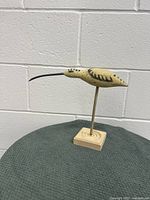 Side view of wooden carved Long-billed Sandpiper figurine mounted on stand against a white brick wall.