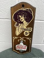 Front view of vintage Coca Cola wooden sign showing graphic of woman, 5 cent price, and mounted metal bottle opener with Coca Cola logo.