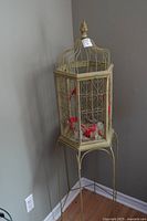 Full view of gold-tone metal bird cage on tall stand against wall