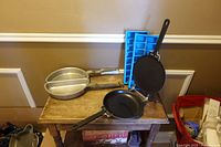 Group view showing aluminum divided skillet, hinged black non-stick pan, and two blue ice cube trays on table