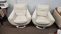 Two white leather swivel armchairs on round stainless steel bases side by side on brown carpet, against a light grey wall.