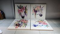 Four 12x12 inch canvas unicorn paintings with floral crowns arranged in a 2x2 grid on the floor, vibrant pastel colors, watercolor style.