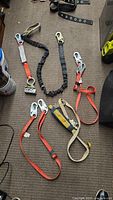 Five safety lanyards laid on carpet featuring various metal hooks and clips, multiple strap colors including red, black, and beige, overall showing signs of wear.