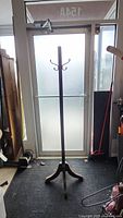 Full view of tall wooden coat rack with tripod base, standing on a black mat in front of a door.
