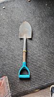 Front view of a small spaded shovel lying on a carpeted floor, showing the metal blade, wooden shaft, and blue plastic handle grip.