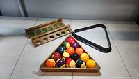 Full set of pool balls arranged in a wooden Brunswick rack with a black plastic triangle rack and a wooden cue rack in the background