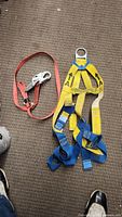 Top view of yellow and blue work harness with metal D-ring and red lanyard with metal snap hook laid on carpet.