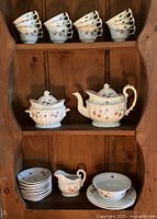 Photo showing four stacks of tea cups on top shelf, teapot and sugar bowl with lids on middle shelf, creamer, small stack of plates and bowl on bottom shelf. Hand-painted strawberry vine decoration visible on all pieces.