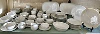 Full view of all the dinnerware pieces spread on a table showing plates, bowls, cups, and serving platters in white porcelain.