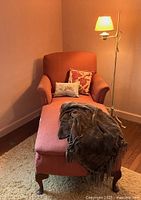 Side angle showing pink chaise lounge with rolled arms, wooden carved legs, two pillows, and chenille throw, against neutral walls and wooden floor