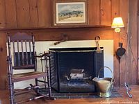 Full view of the fireplace with the rocking chair on the left, fire tools inside the hearth, brass bucket on hearth, folding fire screen, vintage lithograph hung above mantel, and wrought iron floor lamp standing on right.