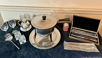 Full lot image showing fondue pot with lid, warming base, tray, forks in case, unopened forks in plastic, sterno can, ladle, candle holders, wine glasses, chrome pitcher with underplate.