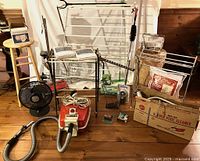Full lot overview showing Eureka vacuum, Hoover Pixie handheld vacuum in box, drying racks, fans, folding grocery cart, wooden stool, and household items.