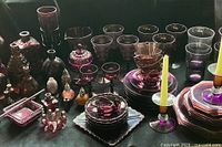 Full view of the amethyst glassware collection showing various glasses, candleholders, vases, plates, and decorative bottles gathered together on a table with a dark cloth.