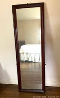 Full front view of tall rectangular mirror with dark mahogany wooden frame, leaning against a wall showing reflection of room including white bed and radiator.
