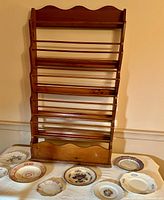 Maple wood magazine rack with scalloped borders standing behind seven displayed plates varied in size and decoration.