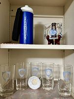 Five Mount Holyoke College emblem tumblers arranged in shelf space, one blue Mount Holyoke water bottle with white lid and strap to left, one small clear glass ashtray to right.