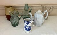 Grouped view of all six pitchers showing variety of materials and designs including stoneware, glass, and transferware ceramics