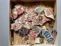 Photo of a box filled with a variety of cancelled postage stamps mostly still attached to small pieces of envelopes. The stamps vary in color including red, blue, green, brown, and purple. Several feature portraits, others have numeric or symbolic designs.