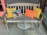 Front view of weathered teak bench with four pillows (two mustard yellow, one orange, one scenic beach design) placed on the seat, showing wooden slats, armrests and curved backrest.