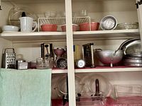 Wide view of shelves showing pink Pyrex mixing bowls, white ceramic pitcher, metal colander, graters, and various glass bakeware.