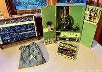 Polaroid Junior microscope in green wooden case with two doors open showing microscope, specimen tubes in racks on doors, drawers with slides, two instruction manuals in front, blue pouch with marbles and button man figure beside.