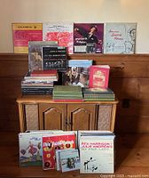 Stacked vinyl LP records, some standing in a row on a cabinet including classical, Broadway, and others, with opera score books displayed.