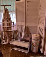 Photo showing collection of vintage pink housewares including three-panel wooden shutter screen on casters, ironing board, luggage rack with strap, turned quilt rack, hand-painted laundry hamper, shoe rack, and perforated metal wall unit in pink pastel finish.