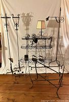 View of all wrought-iron items including two standing candle holders, two lamps, folding table stand, folding three-tiered plant stand, sad irons with stands, wall hangers, and bell