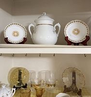 Shelved china plates with gold rims and floral patterns, flanking a white covered pot with handles and gold trim