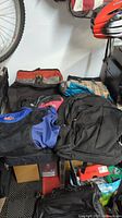 Overview of assorted bags and backpacks piled together, showing different colors and types including insulated lunch bag and black backpacks.