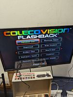 TV screen showing ColecoVision Flashback game menu with game titles such as Alphabet Zoo, Blockade Runner, Aquattack, Brain Strainers, Artillery Duel, Bump'n Jump, Bankruptcy Builder, and Choplifter.