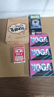 Photo showing all six items: Nasty Things board game, Focus on the Meme game box, three Yoga Smarts game boxes stacked, and 1864 premium giant playing cards.
