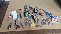 Wide view of all items in the lot spread on table. Includes various bottle openers, keychains, magnifying glasses, scissors, playing cards, and assorted small items.