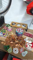 Overview of table with scattered copper decorative pieces, several plastic holiday cookie cutters in red and green, a pink and white floral decorative tin, a box of red plastic kitchen tools, and a large circular copper item.