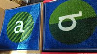 Two carpet squares showing letters 'a' in white on a blue and green striped circle background and 'b' in white on a green and dark green circle background