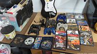 Wide view of controllers and PlayStation games laid on a table including 5 controllers and approximately 15 game cases.