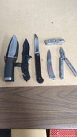 Six knives and multi-tools laid flat on surface: three folding knives, one fixed blade with sheath, small multi-tool, and plier multi-tool