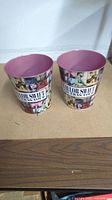 Two metal popcorn buckets decorated with Taylor Swift The Eras Tour images and text, with pink interior, shown side by side on a wooden surface.