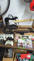 Six Xbox 360 wireless controllers in white and black, two guitar controllers for Guitar Hero or similar game, games and Kinect sensor arranged on table.