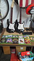 Two black and white Guitar Hero imitation Fender guitar controllers against a wall, Xbox One Kinect sensor at side, various Xbox One game cases laid out on table.