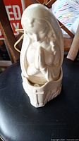 Photo of white ceramic Blessed Mother figurine with praying hands, shown on a black chair with visible power cord at the back.