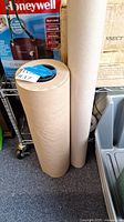 Photo of two large rolls of brown kraft paper standing upright on a floor, with packaging boxes behind them.
