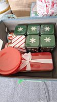 Top view of a box containing assorted holiday-themed tins including four green small square tins with snowflake designs, a candy cane striped rectangular tin, and a red rectangular tin with a white ribbon print.