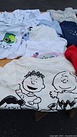 Photo of multiple girls shirts including Peanuts characters shirt and white collared shirts laid out on a wooden board